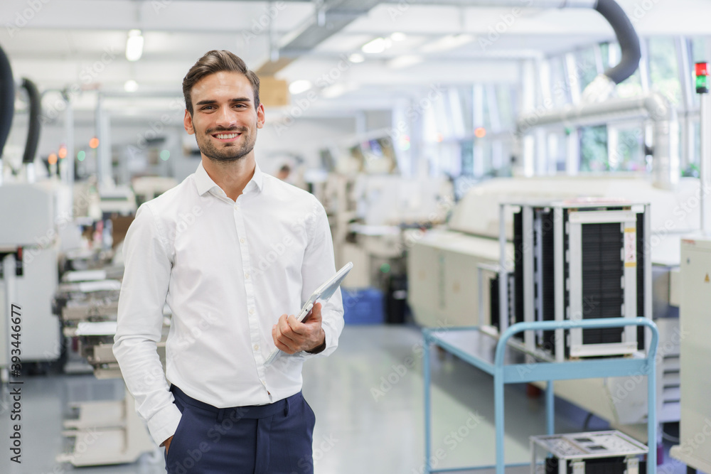 Smiling confident young businessman holding digital tablet while standing with hands in pockets at illuminated factory Smiling confident young businessman holding digital tablet while standing with hands in pockets at illuminated factory