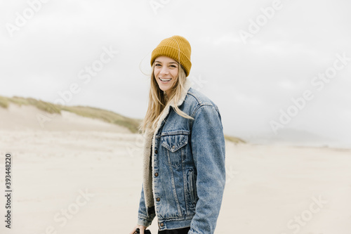 young woman wearing yellow beanie and denim jacket on empty beach smiling toward camera