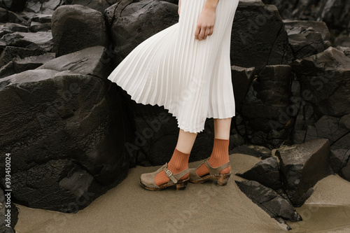 young person wearing flowing white skirt and orange socks standing in front of dark rocks