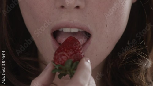 Close-up european positive woman eating ripe delicious red strawberry on sparkles background in cafe. Health and nutrition concept. Slow motion