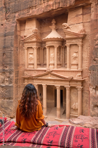 A girl in an orange shirt sits on the edge of a cliff on a carpet and looks at the ancient city of Petra