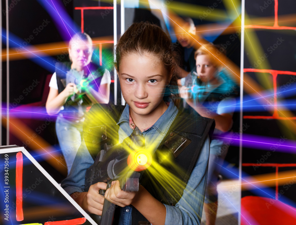 Cheerful girl aiming laser gun at other players during laser tag game in dark room