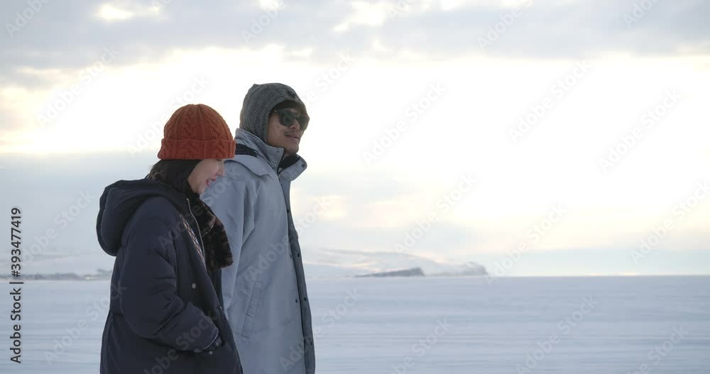 Young asian loving couple walking together in the distance on the ice of a frozen snow covered endless winter lake in slow motion. Date. Valentines Day in winter at sunset. Love and Romance Theme. Young asian loving couple walking together in the distance on the ice of a frozen snow covered endless winter lake in slow motion. Date. Valentines Day in winter at sunset. Love and Romance Theme.