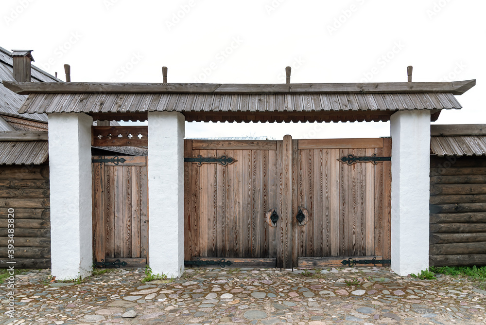 Old weathered wooden gate with small door and green roof at fence made with large grey logs near traditional rural house under blue sky on sunny, summer day Old weathered wooden gate with small door and green roof at fence made with large grey logs near traditional rural house under blue sky on sunny, summer day