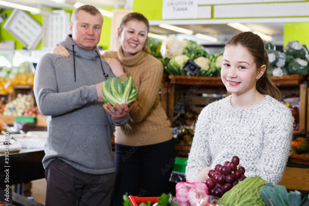Smiling preteen girl choosing ripe fruits and vegetables while shopping with parents in greengrocery