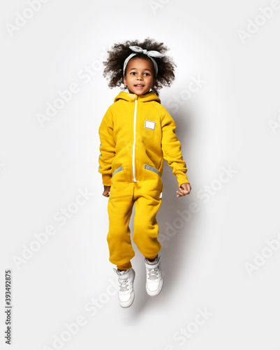 Happy sporty dark-skinned curly kid girl in warm comfortable yellow sport jumpsuit does fitness exercises jumps, acrobatics, aerobics, gymnastics in sports school