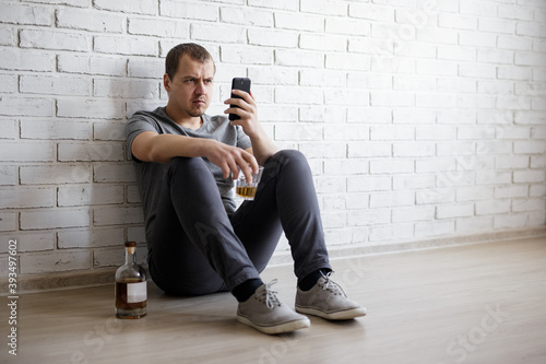 alcoholism and depression concept - young man sitting on the floor, drinking alcohol and using smartphone
