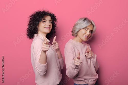 Cute curly women in pink sweaters are pointing at camera and smile on a red studio wall
