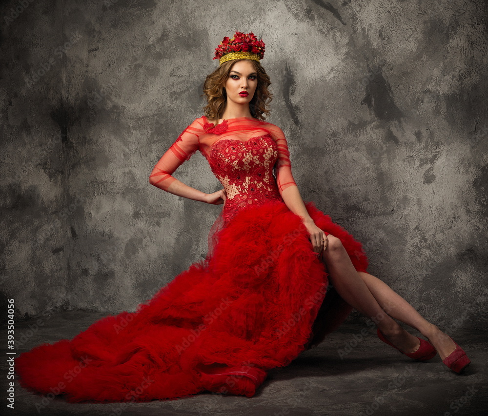 beautiful slender model in a red stylish dress with a crown of living roses on her head sits against an old wall