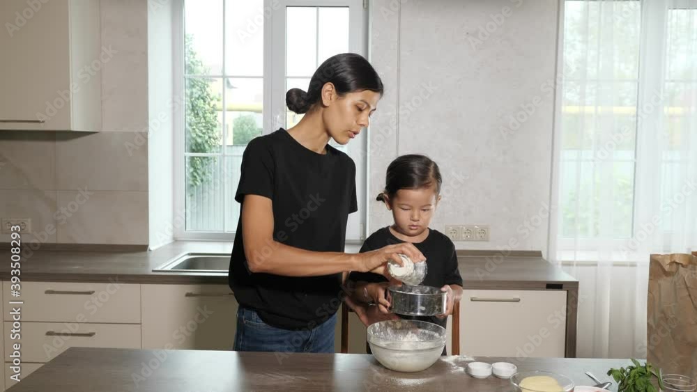 Beautiful mother pours flour from cup and little daughter wearing black shirts and same hairstyle shakes metal sieve above glass bowl Beautiful mother pours flour from cup and little daughter wearing black shirts and same hairstyle shakes metal sieve above glass bowl