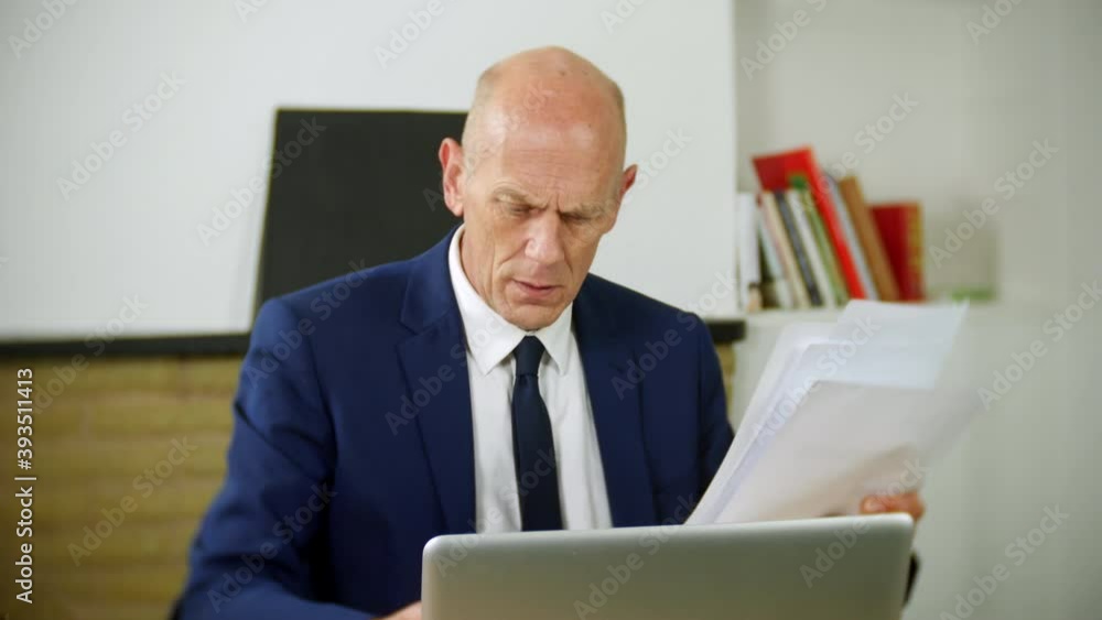 A mature businessman is confused and frustrated with working from home remotely. He is looking at paperwork while using a laptop computer. A mature businessman is confused and frustrated with working from home remotely. He is looking at paperwork while using a laptop computer.