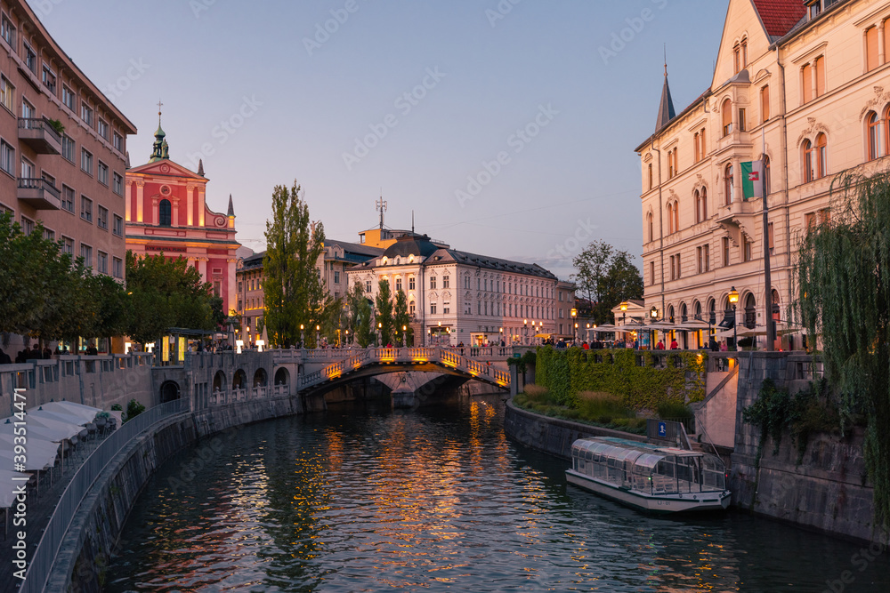 The promenade on the Ljubljanica river in the slovenian capital city Ljubljana with the Triple bridge and famous pink franciscan church in the evening