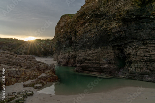 sun peaks over the edge of a cliff with sandy beach and tidal pools in foreground