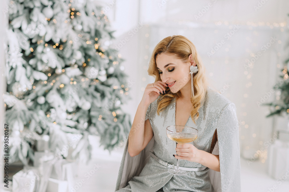 Smiling beautiful blonde woman in a luxurious outfit with a glass in her hands against the background of blurry lights bokeh of a Christmas tree. Happy New Year 2021. Soft selective focus, art noise. Smiling beautiful blonde woman in a luxurious outfit with a glass in her hands against the background of blurry lights bokeh of a Christmas tree. Happy New Year 2021. Soft selective focus, art noise.