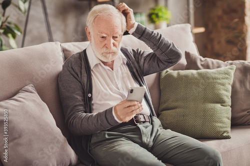 Photo of cute thoughtful pensioner wear grey cardigan sitting couch holding modern gadget indoors house flat