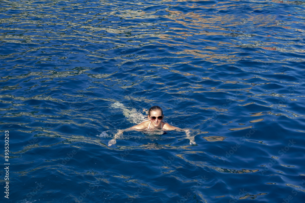 Young adult woman in sunglasses swims in blue water. Cute caucasian girl bathes in the pool, sea, river, lake, etc.