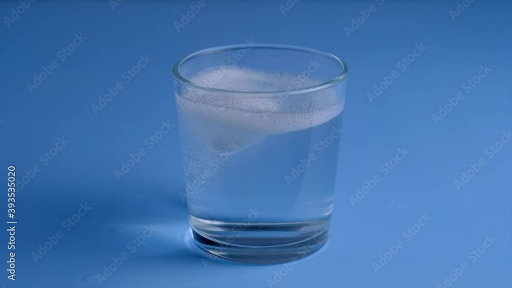 Effervescent white tablet falls in glass on blue background. Fizzy tablet. Slow motion. Effervescent white tablet falls in glass on blue background. Fizzy tablet. Slow motion.