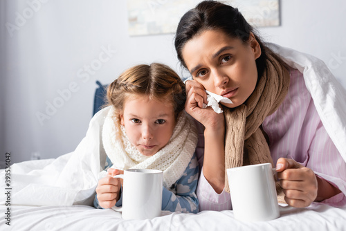 sick, upset woman and daughter looking at camera while lying in bed with warm drink
