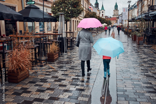 Back view of mother and her daughter holding the pink and blue umbrellas walking in a downtown on rainy gloomy autumn day