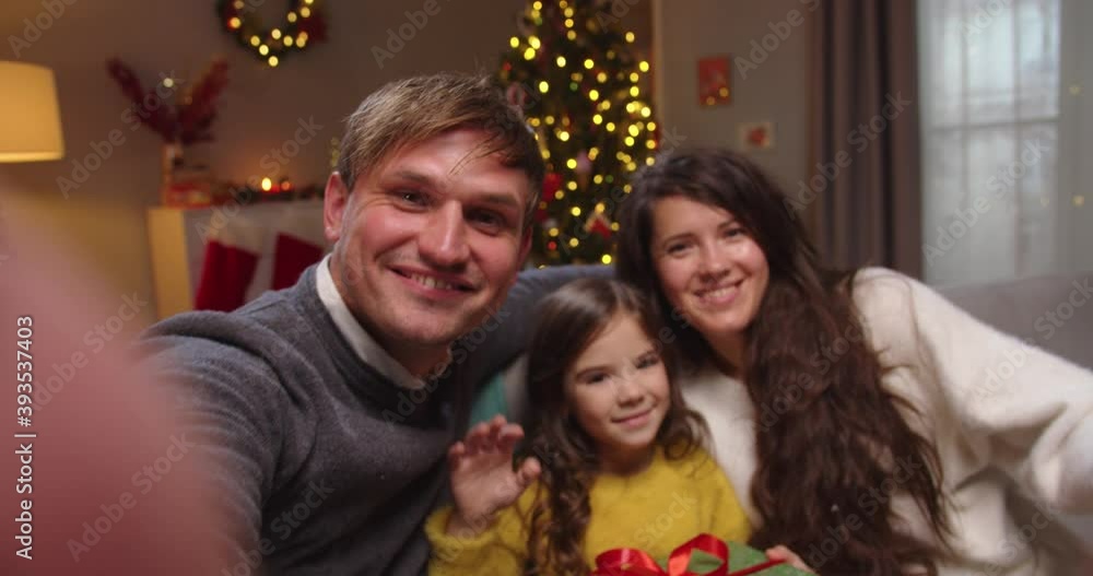 Close up. POV of joyful Caucasian young family with little daughter smiling and waving hands while videochatting online in decorated room with glowing xmas tree on New Year Winter holidays celebration Close up. POV of joyful Caucasian young family with little daughter smiling and waving hands while videochatting online in decorated room with glowing xmas tree on New Year Winter holidays celebration