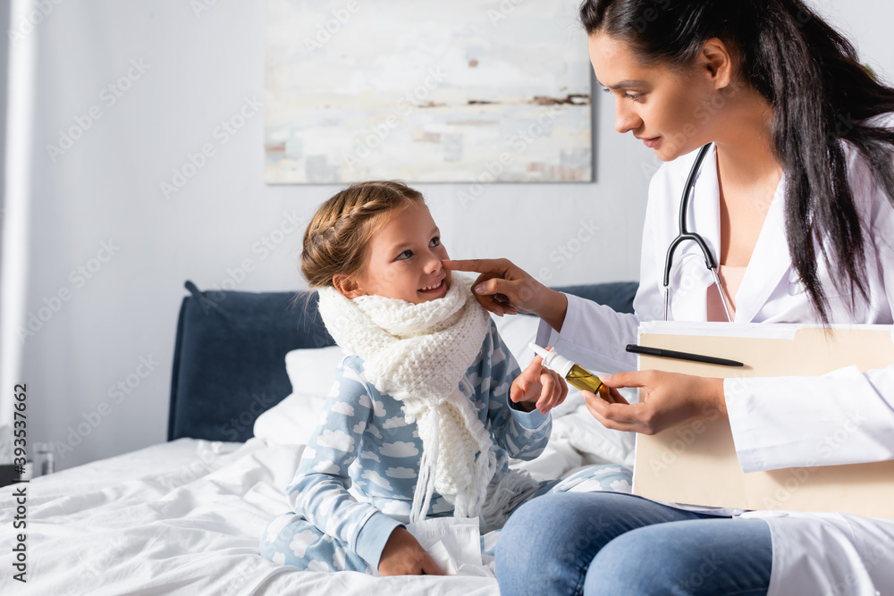 pediatrician holding nasal spray while touching nose of smiling girl in warm scarf
