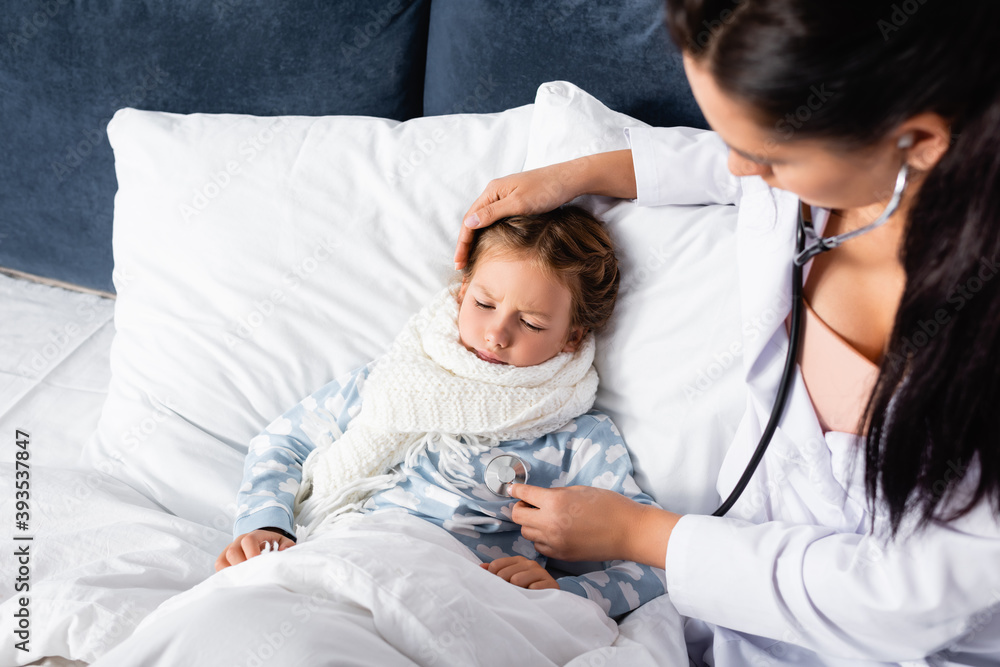 high angle view of pediatrician touching head of sick girl and examining her with stethoscope on blurred foreground