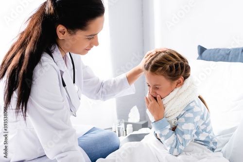 doctor touching head of squeezing girl sitting in bed