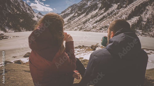 Couple drinking hot coffee or tea and sitting next to stunning winter landscape. Concept of travel and adventure at the frozen Gaube Lake (Lac de Gaube) in the French Pyrenees, France.