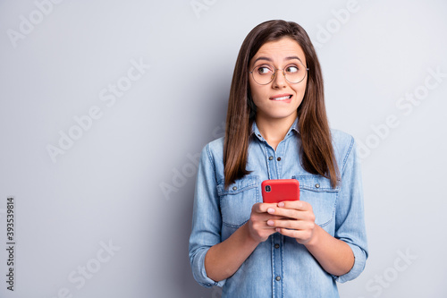 Photo of doubtful charming young woman look empty space hesitate hold phone wear denim shirt isolated on grey color background