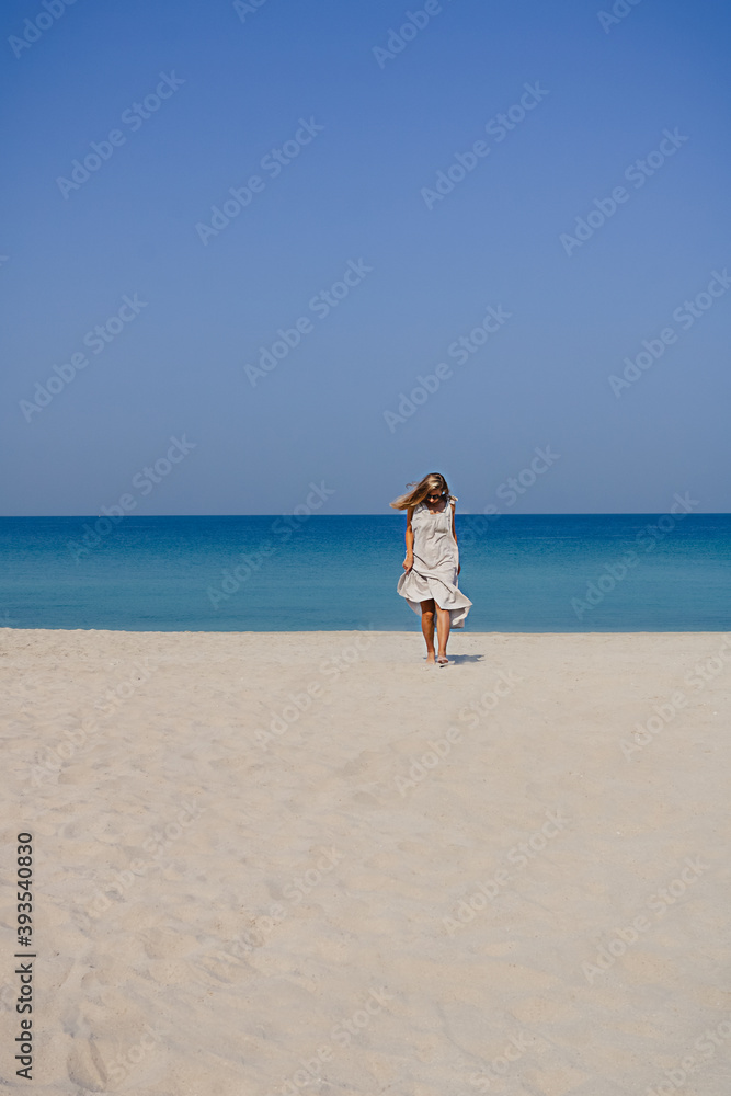 A blonde smiling girl in a maxi linen dress with fluttering hair jumping and dancing on a sand beach against blue sky and sea background A blonde smiling girl in a maxi linen dress with fluttering hair jumping and dancing on a sand beach against blue sky and sea background