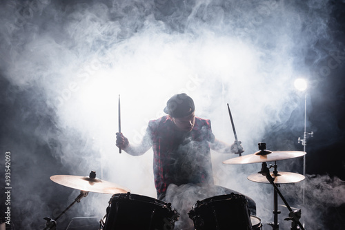 Musician with drumsticks playing while sitting at drum kit with smoke and backlit on background