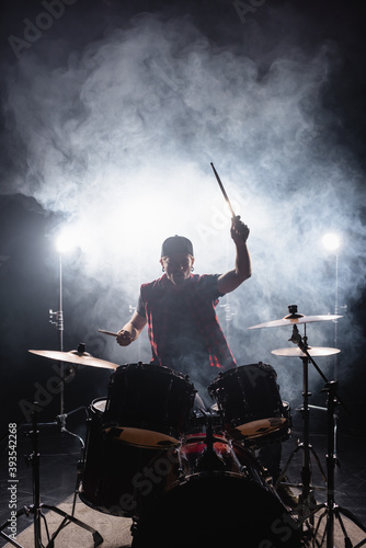 Fotomurale Man playing drums while sitting at drum kit with backlit and smoke on background