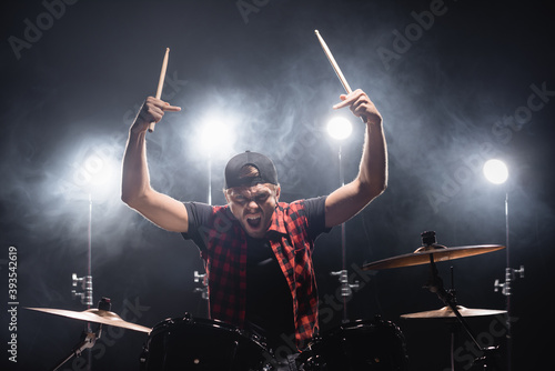 Aggressive drummer with middle fingers looking at camera and holding drumsticks near drum kit with backlit on background