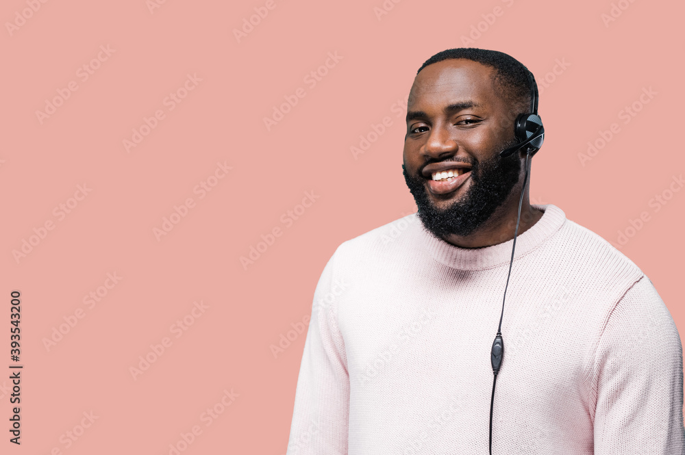 Fototapeta premium Close-up portrait of young african american man worker of a call-centre with headphones standing on a pink isolated background looking at the camera and smiling