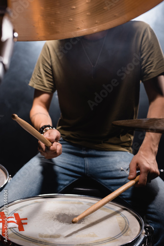 Obscure face of musician with drumsticks playing drum on black