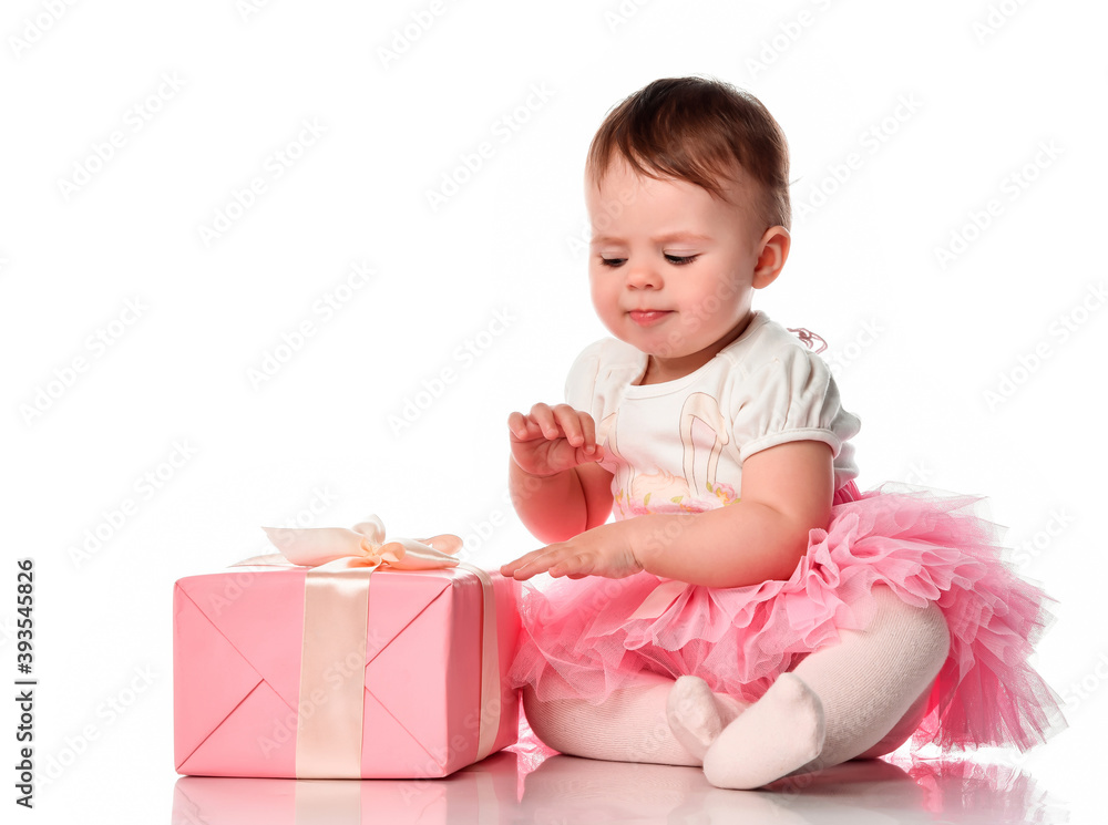 Mistrustful shy cute baby girl sitting on white floor background and looking at gift box studio shot full length portrait. Congratulation and surprise for excited curious beautiful toddler child Mistrustful shy cute baby girl sitting on white floor background and looking at gift box studio shot full length portrait. Congratulation and surprise for excited curious beautiful toddler child