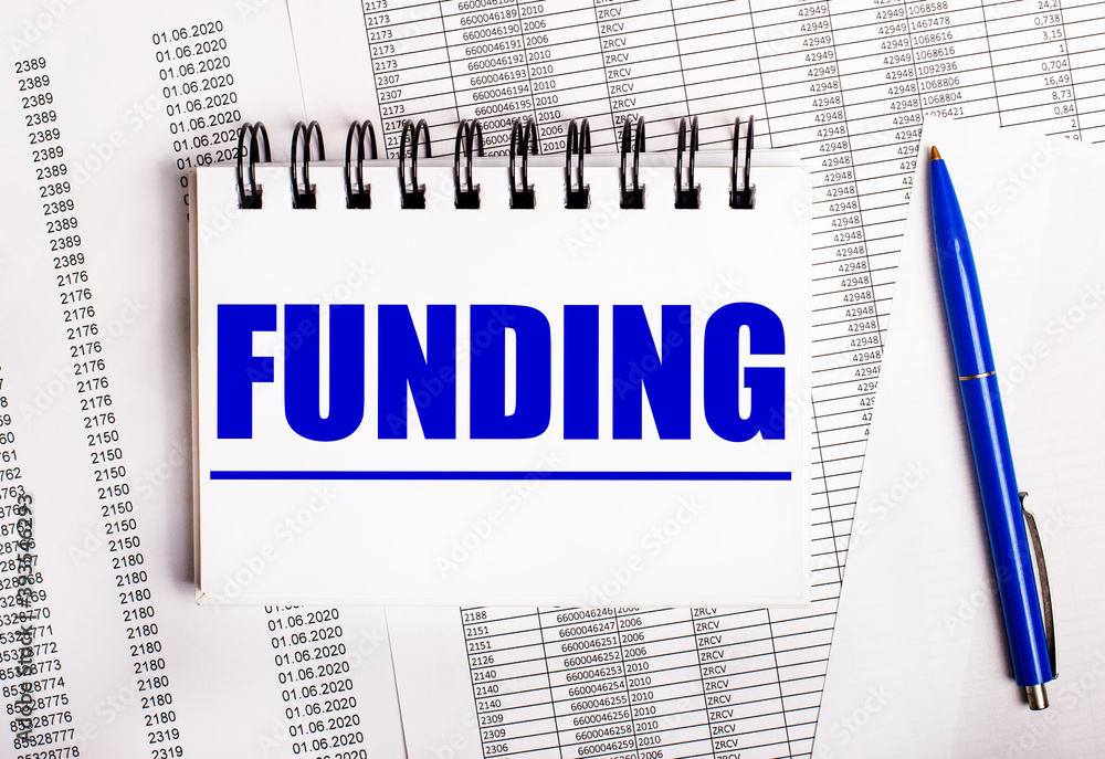 On the table are charts and reports, on which lie a blue pen and a notebook with the word FUNDING On the table are charts and reports, on which lie a blue pen and a notebook with the word FUNDING