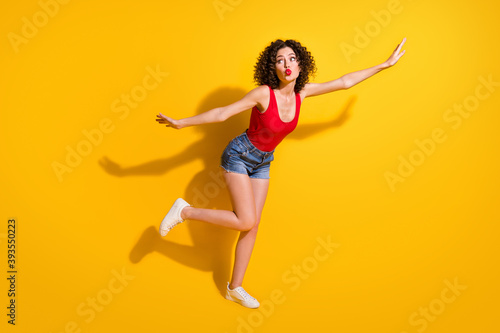 Full size photo of lovely dreamy young girl raise hands leg lips send air kiss attract guys party wear red singlet unclothed shoulders denim shorts isolated vibrant yellow color background