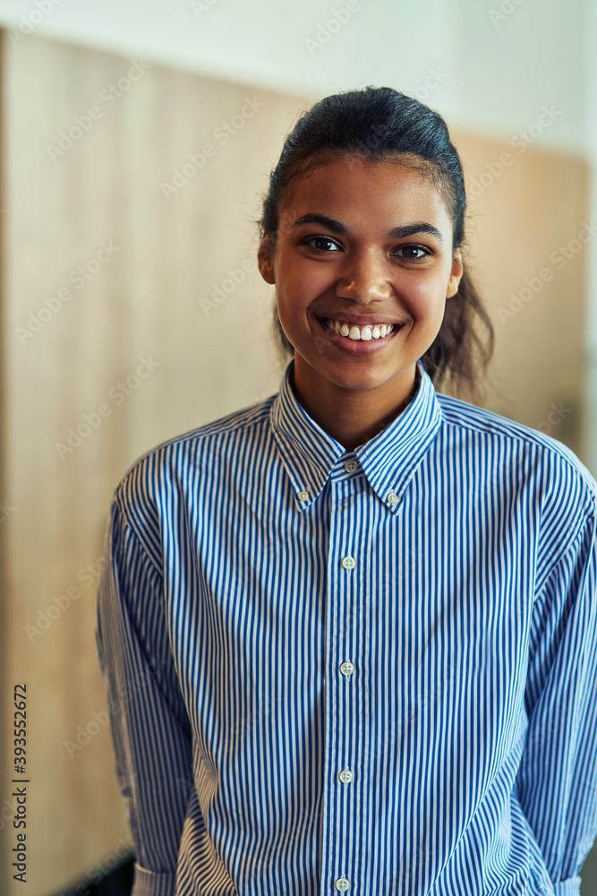 Vertical shot of a young happy mixed race business woman in blue shirt looking at camera and smiling while posing in the office
