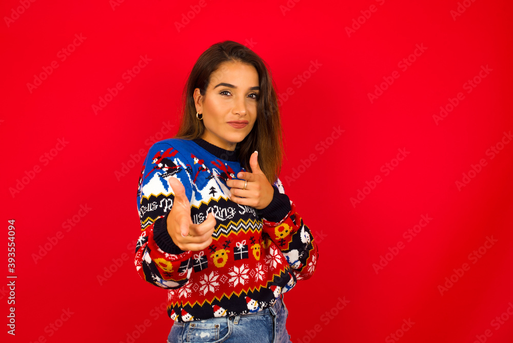 Hey you, bang. Joyful and charismatic good-looking young beautiful Caucasian woman wearing Christmas sweater against red wall,  winking and pointing with finger pistols at camera happily and cheeky.