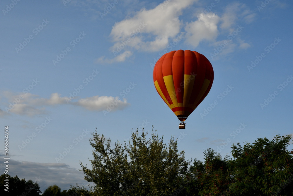 Close up on a red and yellow balloon with a big basket underneath flying through the cloudy sky full of clouds next to a dense forest or moor seen on a sunny summer day on a Polish countryside