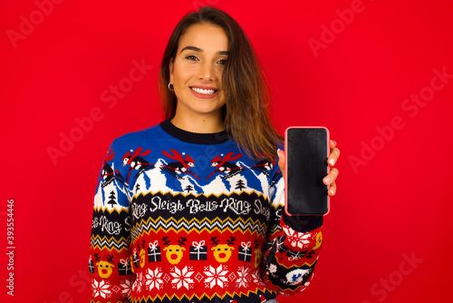 Smiling young beautiful Caucasian woman wearing Christmas sweater against red wall, showing empty phone screen. Advertisement and communication concept.