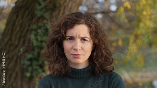Portrait of beautiful confused, angry or annoyed young woman in nature. Attractive frowning millennial girl with curly hair at park or forest during sunset looking at camera. Slow-motion.