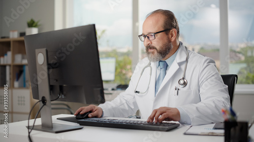 Hispanic Doctor's Office: Experienced Physician Sitting at His Desk Working on Personal Computer. Health Care Specialist Filling Medicine Prescription Documents, Checking Analysis Test Results.