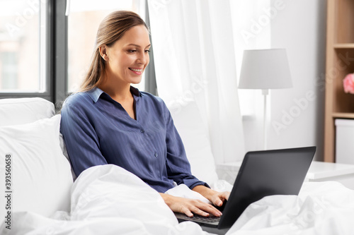 technology, internet and people concept - happy smiling young woman with laptop computer in bed at home bedroom