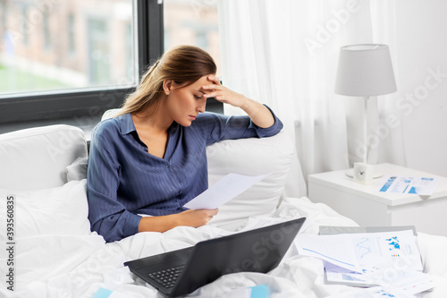 remote job  people and business concept - stressed young woman with laptop computer and papers in bed working at home