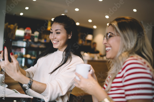 Smiling multiracial female best friends enjoying meeting in cafe for coffee making selfie on smartphone camera, positive woman share content on social networks spending time with happy colleague