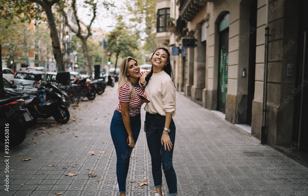 Full length portrait of cheerful diverse tourists laughing at camera during walking time at urban setting, happy hipster girls with cellphone gadget satisfied with sincerely friendship relations