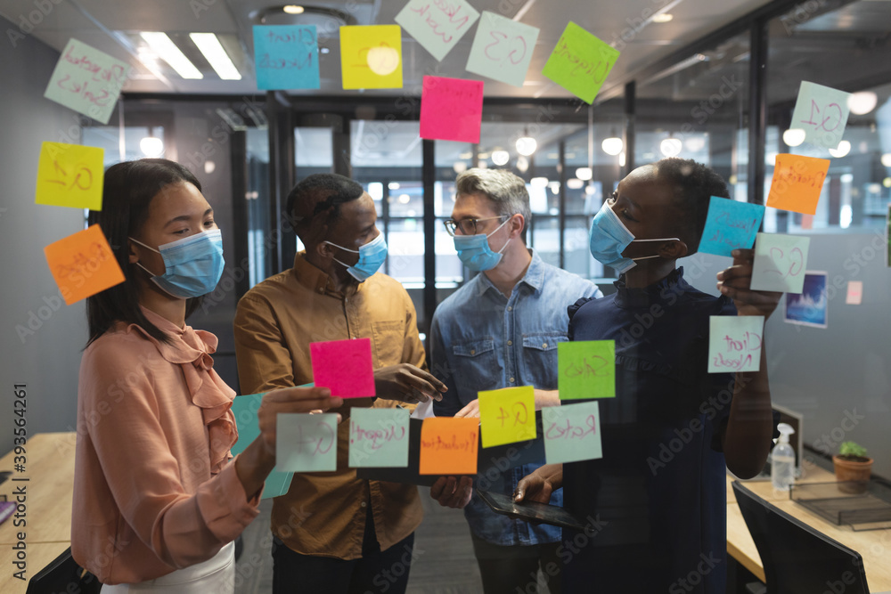 © Wavebreak Media - Diverse office colleagues wearing face masks discussing over memo notes on glass board at modern off