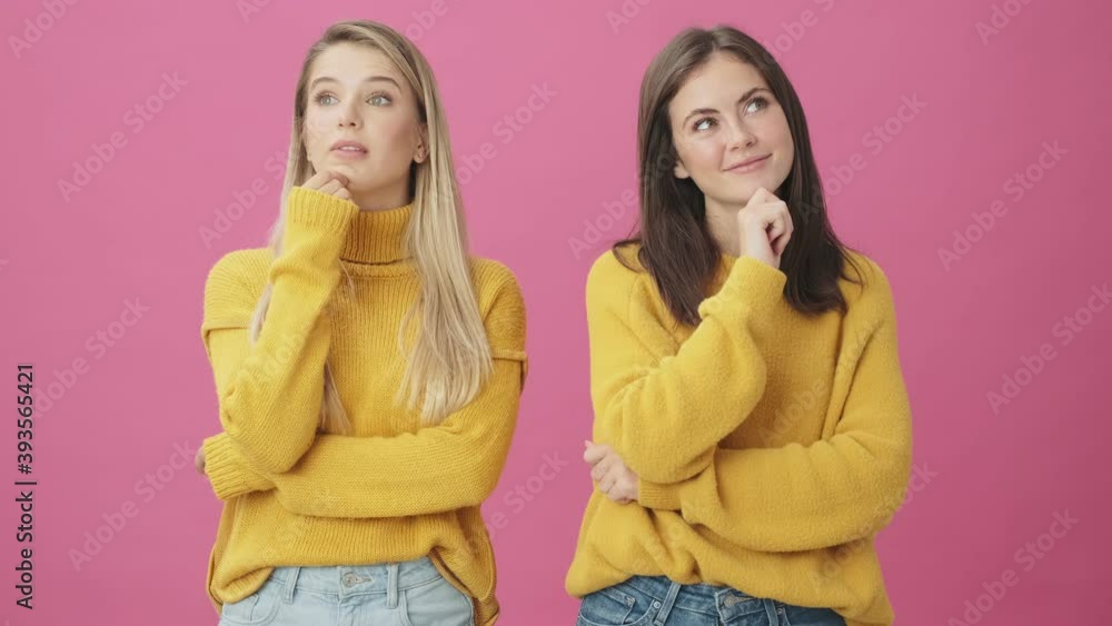 Two thinking women wearing yellow sweaters are coming-up with an idea while raising fingers standing isolated over pink background in studio Two thinking women wearing yellow sweaters are coming-up with an idea while raising fingers standing isolated over pink background in studio